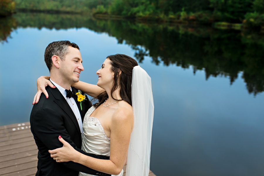 A bride and groom hold each other on the dock of a lake with the reflection of the sky in the water.